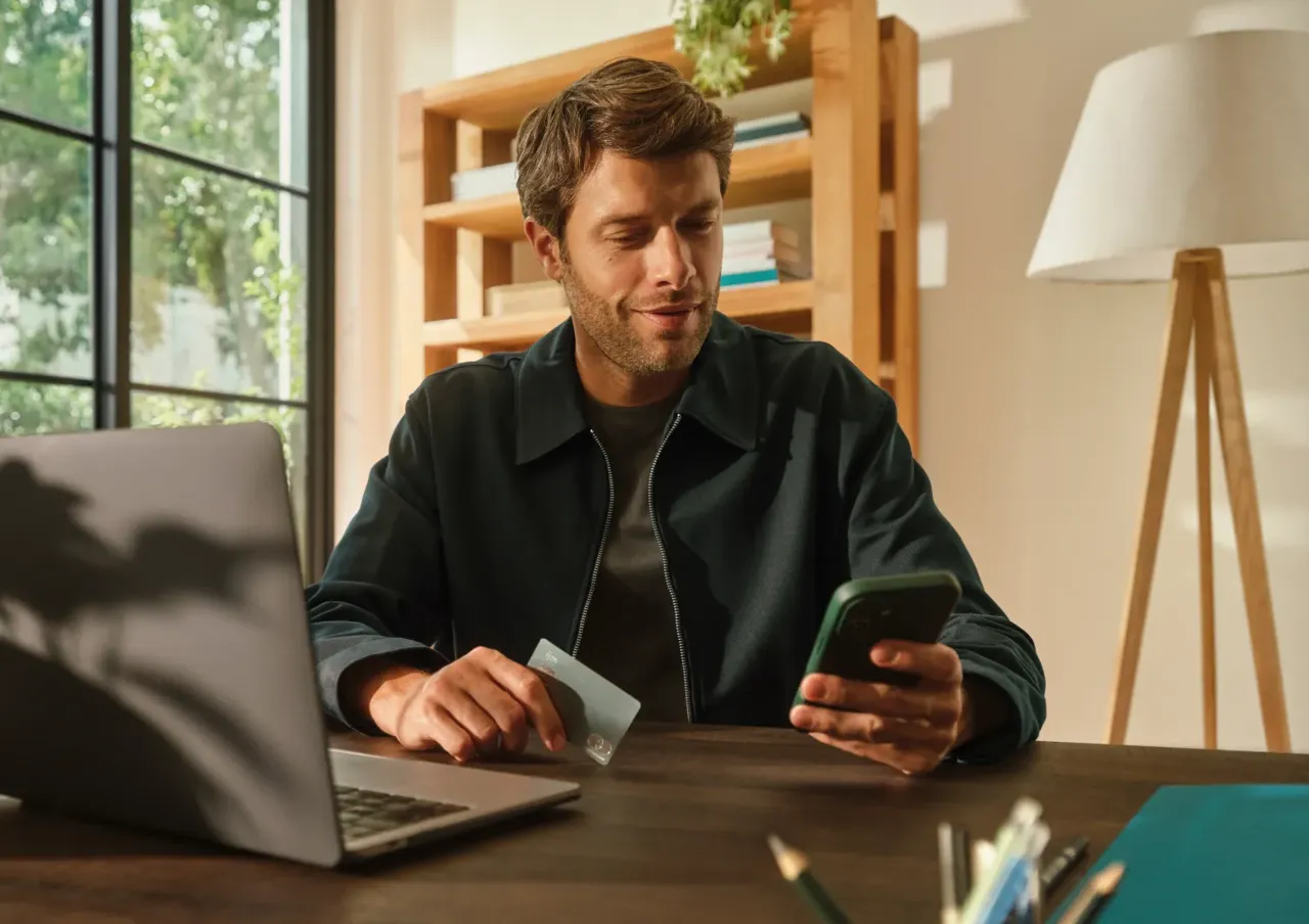A man sits at a desk with an open laptop, holding a payment card while looking at his smartphone screen.